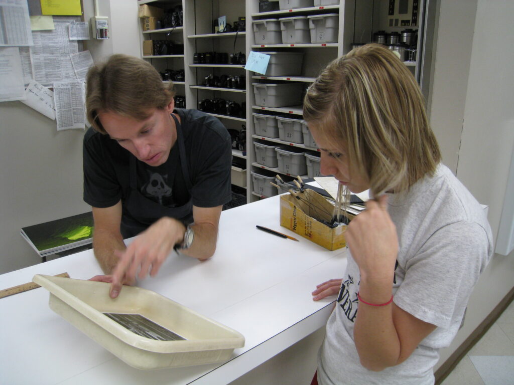 Photography Academic Lab Technician Mark Magavern goes over exposure and contrast with a student working in the black and white darkroom. Students used to expose their images and bring them out into the light to get direction on what to do to improve them. Archive photo.