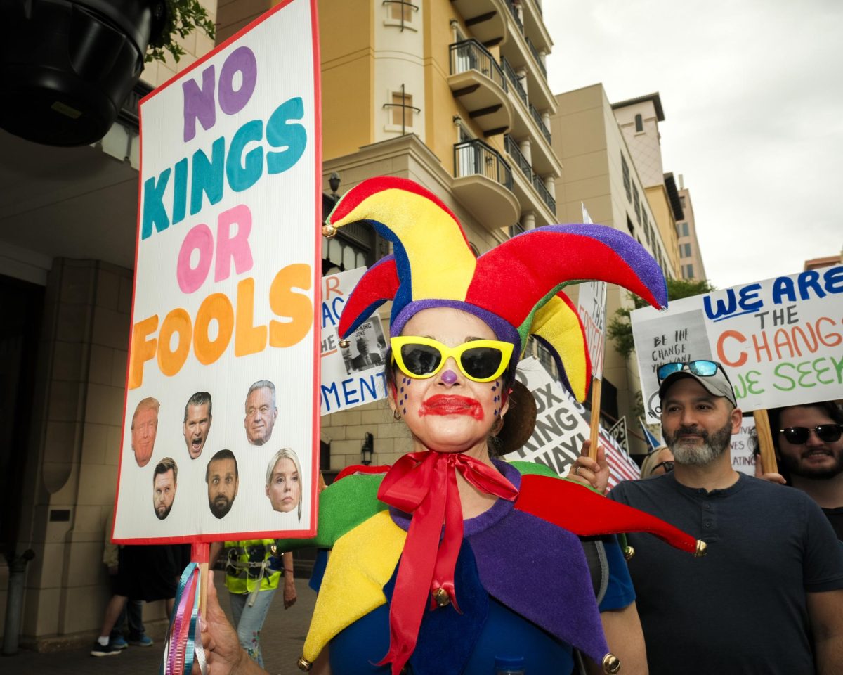 A Protester dressed like a court jester holds a sign saying "No Kings or Fools" with photos of prominent members of President Trump's Cabinet at the No Kings Protest on Saturday.
